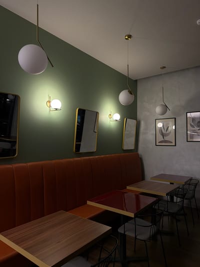 The media file displays the interior of a modern and stylish restaurant or cafe in Florești, Moldova, viewed from a tilted perspective, where the long axis of the room appears to run diagonally. The space features a continuous, reddish-brown padded booth, likely intended for multiple diners. In front of the booth are several small, square tables; some have warm wooden tops, while others feature a reflective, dark red surface, all bordered by a light-colored trim. Opposite these tables, a series of black wire-frame chairs are arranged.
The room's walls are distinctly two-toned: the lower section is painted a muted olive green, and the upper section, extending onto the ceiling, is a light, textured grey. Contemporary lighting fixtures illuminate the space. Several large, spherical white globe lights are suspended on slender, brass-colored arms, some appearing to extend from the ceiling/upper wall and others from the green wall. Smaller, spherical wall sconces with brass-colored bases are also mounted on the green wall, casting bright, localized light.
Decorative elements include multiple rectangular mirrors with thin brass frames on the green wall, and two framed abstract art pieces in muted tones on the grey wall. A white smoke detector or similar device is visible on the ceiling. The artificial lighting suggests an evening setting or an intentionally curated ambiance. No people are present, indicating an empty dining area. The scene conveys a chic, inviting, and calm atmosphere. No visible text is present.