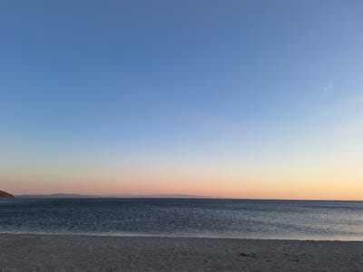 A wide-angle, low-angle shot captures a serene seascape at dawn or dusk in Erdek, Turkey. The foreground is dominated by a sandy beach, appearing light gray and textured with subtle footprints. The midground features a calm, dark blue sea, its surface rippling gently. In the distance, a low-lying landmass is visible, its silhouette a soft, muted brown against the horizon. The sky transitions from a deep blue at the top to a pale orange and pink near the horizon, suggesting the sun is either rising or setting. The scene is devoid of any people or structures, emphasizing the natural beauty of the coastal environment.