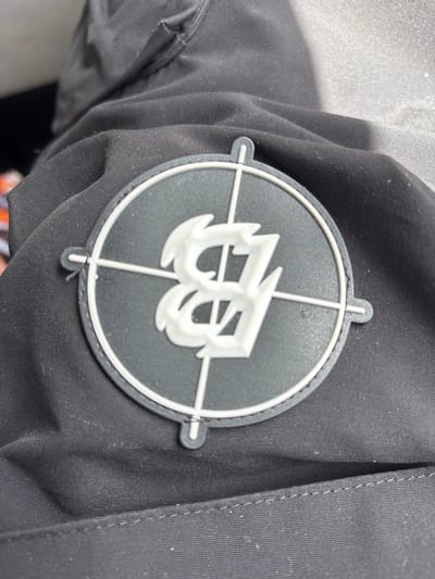 The image shows a black jacket with a circular patch on the upper arm. The patch is black with a white border and crosshairs. A stylized white letter "B" is superimposed over the crosshairs. The jacket appears to be made of a dark, possibly synthetic, fabric. There is no visible indication of the location being Philadelphia.