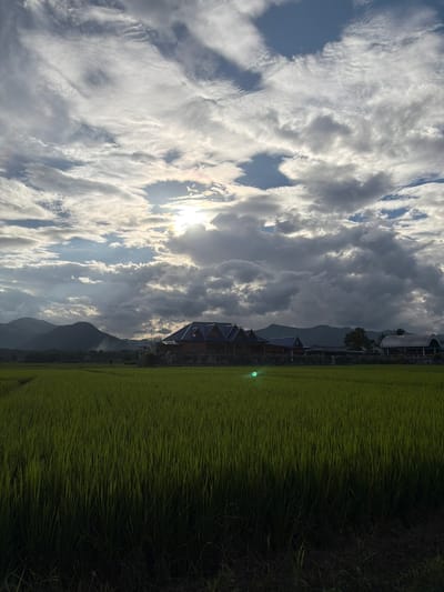 The photograph depicts a lush, green rice field in the foreground, stretching towards a line of silhouetted mountains in the distance. A large building with a blue roof and traditional architectural elements is situated in the mid-ground, to the right of the mountains. The sky above is filled with dramatic, textured clouds, with the sun peeking through in the upper center, casting rays of light and a subtle rainbow effect. A small, bright green circular object is visible in the rice field, approximately one-third of the way from the left edge of the frame, seemingly reflecting light. The scene appears to be an outdoor landscape, possibly in the rural outskirts of Ban Pong, Thailand, characterized by agricultural land and natural topography under a dynamic sky. There are no people or animals visible in the frame.