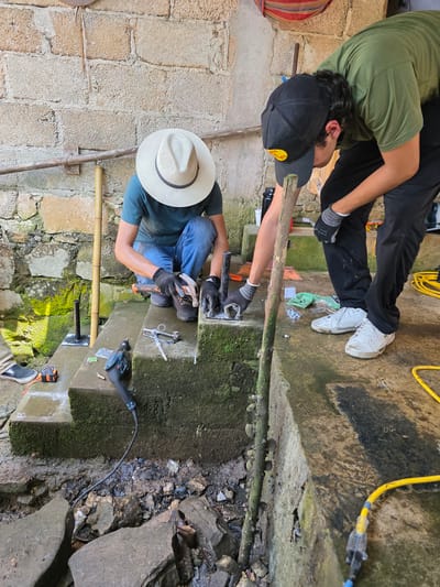 Two men are engaged in construction work outdoors on stone stairs in Pinahuista, Mexico. One man wears a light-colored hat, a blue-green shirt, and jeans. He is kneeling, hammering a metal object with a small hammer. Nearby are wrenches, a drill, and a tape measure. The second man wears a green shirt, black pants, a black baseball cap, and work gloves. He is holding a long wooden beam in place. The steps are covered in moss and algae. A yellow extension cord is visible in the background. The environment suggests a rustic or rural setting.