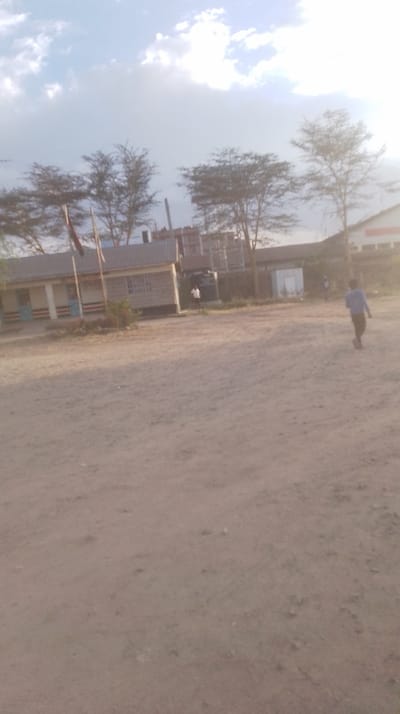 A dirt field dominates the foreground. In the background are low buildings and trees under a bright, partly cloudy sky. There is a flag pole on the left, next to a building with red and blue stripes. A man in a white shirt is standing near a building with a water tank. A person in a blue shirt and dark pants walks across the field. The image appears to be taken in a rural, sunny area.