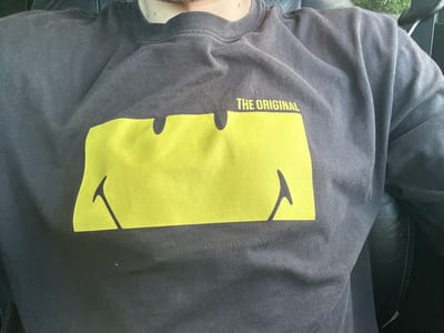 A person is wearing a dark gray crewneck sweatshirt with a large yellow graphic on the front. The graphic is a stylized smiley face with a wide mouth that resembles the Batman logo. Above the graphic, the words "THE ORIGINAL" are printed in yellow text. The person's chest, neck, and shoulders are visible. The sweatshirt appears to be made of cotton and has a slightly textured surface. The background is dark and appears to be indoors, possibly a car seat due to the texture and stitching visible on the right side of the image. There is no discernible information related to Shatava, Ukraine.