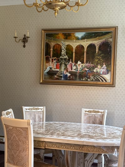 The image shows an indoor dining room. The walls are covered in a pale, textured wallpaper with a subtle repeating pattern. A large, ornate framed painting hangs on the wall, depicting a scene with classical figures in a garden setting by a fountain. Above the painting, a brass chandelier with multiple arms is visible. To the left of the painting, a brass wall sconce with two candle-like lights is mounted on the wall. In the foreground, a large oval dining table with a marble-patterned top is covered with a clear plastic protective sheet. The table has ornate carved details and is surrounded by several upholstered dining chairs with decorative patterns in gold and cream. The chairs have high backs with golden ornate trim and upholstered seats. The scene appears to be a well-decorated dining area, possibly in a home.