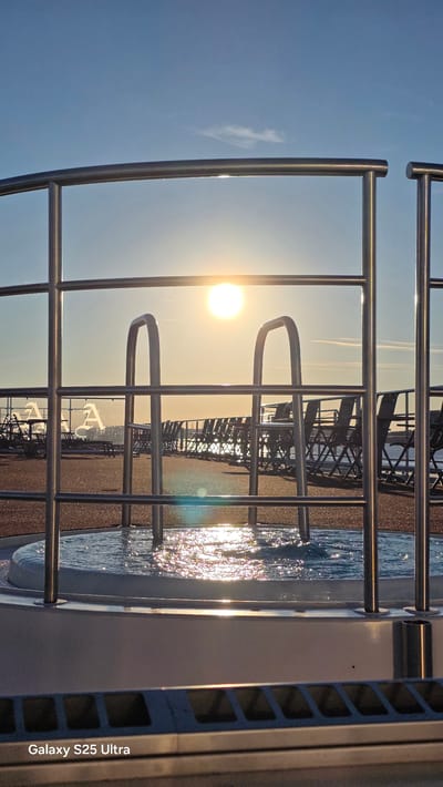 The image depicts a close-up view of a hot tub on a deck, possibly a cruise ship or hotel, during sunset. The sun is low in the sky, casting a warm glow and creating reflections on the water's surface. Metal railings and a ladder for the hot tub are prominent in the foreground, framing the scene. In the background, rows of empty deck chairs are visible, suggesting a tranquil atmosphere. The ocean or a large body of water stretches to the horizon, meeting a clear blue sky with a few wispy clouds. The overall impression is one of relaxation and a serene end to the day. There is no discernible location in the city of Giurgiu, Romania mentioned or depicted in the image.