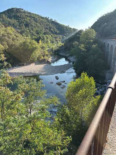 The image captures a scenic view of a river flowing through a valley, with a stone bridge arches visible on the right side of the frame. The river is surrounded by lush green trees and vegetation covering the steep hillsides. The sun is shining, casting a warm glow on the landscape, with some light rays piercing through the trees in the background. A gravelly riverbed is visible along the banks, with smooth stones scattered in and around the water. The bridge, constructed of stone, features multiple arches and appears to be an old structure, possibly part of a railway line given its design. The foreground is partially obstructed by a metal railing, suggesting the photo was taken from a vantage point, likely a bridge or a walkway. The overall impression is one of natural beauty and historical architecture harmoniously coexisting. The location is identified as Générargues, France, a commune in the Gard department.