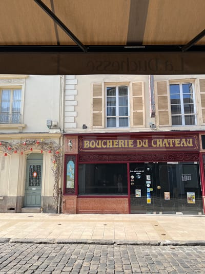 The image shows the facade of a building with two storefronts on a street in Rambouillet, France. The storefront on the right is a butcher shop, identified by the sign "BOUCHERIE DU CHATEAU" in gold lettering on a dark red background. The shop has large glass windows displaying its interior and features a telephone number, "(01.34.83.00.37)", and opening hours posted on the glass. A decorative vine with red bell-shaped ornaments is draped across the facade above the entrance to the left storefront, which is a light green door with a decorative metal gate. The building has an awning extending over the storefronts, casting a shadow. The street in front of the building is paved with cobblestones and a lighter tiled walkway.