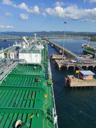 The image depicts a large, green-hulled tanker ship docked at an industrial pier under a bright, sunny sky with scattered white clouds. The scene is set on calm blue waters, with a coastline featuring mountains and a coastal settlement or industrial area in the background.

The primary subject is the large vessel, which appears to be an LNG or LPG carrier, identifiable by its distinctive white structures, extensive network of pipes, and numerous valves on its deck. The ship is moored alongside a concrete jetty, with loading arms and pipelines connected, indicating ongoing loading or unloading operations. Yellow safety markings are visible on the ship's deck and hull.

The concrete pier extends from the shore, supporting a long pipeline bridge that transports materials to and from the land-based facility. A small building with a light blue roof is situated on the pier, likely serving as a control room or office. Another, smaller vessel is visible docked further along the pier in the distance.

The time of day is clearly daylight, with good visibility and bright sunlight reflecting off the water. The weather is fair, with a clear sky. A notable detail on the ship's deck is a person wearing what appears to be a white protective suit or hazmat suit, suggesting the handling of potentially hazardous materials.

Visible text on the ship includes "NO SMOKING" and "SAFETY FIRST" on a white sign, "FLAT ACCESS POINT," and "KEEP CLEAR" on the green hull. Other markings like "WB7S" and "OPEN" on a manhole cover are also visible. The location of this industrial maritime activity is Kunak, Malaysia.