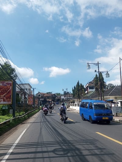 This image captures a street scene in the city of Naung Bo Oe, Myanmar, under a clear blue sky with scattered white cumulus clouds. The perspective is from the road, looking forward down a paved two-lane asphalt road with a solid white line on the left shoulder and a double yellow line delineating lanes. Overhead utility wires cast prominent shadows across the roadway, indicative of bright sunlight.

On the left side of the road, a green grassy embankment leads to a metal guardrail. Beyond this, a large advertising billboard displays a close-up image of a breaded food item, with text "200 M" visible at the bottom. Further down, several multi-story buildings of varying architectural styles are visible behind dense foliage and utility poles.

On the right side of the road, a concrete sidewalk is bordered by low white decorative barriers. Multiple utility poles support ornate, lantern-style street lamps. Tall, slender conifer-like trees are interspersed among more multi-story buildings, one of which features a distinctive green-tiled roof.

Traffic consists primarily of motorcycles and one blue passenger van. In the foreground, a motorcyclist wearing a light-colored shirt, dark shorts, and a helmet, with a backpack, operates a dark-colored motorcycle, moving away from the viewer. Several other motorcycles, some with passengers, are visible further down the road, also traveling in the same direction. A blue passenger van, bearing the license plate "F 1967 ZD", is positioned on the right side of the road, facing away from the viewer. No direct interactions between individuals are discernible.