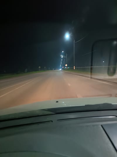 The image is a nighttime, first-person perspective shot from inside a vehicle, looking forward through the windshield. The vehicle appears to be on a road with multiple lanes, indicated by white lane markings. Streetlights line the right side of the road, illuminating the asphalt and the surrounding dark environment. The road stretches into the distance, where the lights become more diffused due to atmospheric conditions. The dashboard of the vehicle is visible in the foreground, partially obscuring the lower part of the road. The environment suggests a rural or suburban setting outside of a densely populated urban center, given the presence of streetlights along a road with open land beside it. No people or other vehicles are clearly identifiable in the scene. The overall impression is one of a nighttime drive through an unpopulated area.
