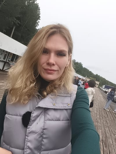 The image is a selfie taken by a young adult female, positioned slightly off-center to the left, standing outdoors on a wooden boardwalk in Nizhnie Yaki, Russia. She has long, wavy blonde hair and is looking directly at the camera with a neutral expression, exhibiting a slight smile. She wears a dark green ribbed long-sleeved top beneath a light grey puffer vest. A small black fuzzy object is affixed to the left side of her vest. Her visible left ear features a small gold hoop earring.
To her upper left, partially visible, is a white building or kiosk with dark Cyrillic lettering, framed by a dense stand of green trees under an overcast sky. The wooden boardwalk, composed of planks, extends into the distance on the right side of the frame. Multiple individuals populate the midground and background. A person with short dark hair, wearing a light-colored hooded jacket and carrying a black bag, is seen from the back, facing away from the camera. Further down the boardwalk, a group of people congregates near a light-colored building with dark, possibly Cyrillic, text on its facade (appears reversed, possibly from a reflection or signage style). A row of several white cars, followed by a white and blue truck, is parked parallel to the boardwalk. A line of trees forms the horizon beyond the parked vehicles. The sky is uniformly overcast, indicating a cloudy day.