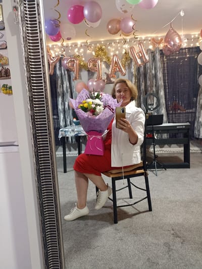 A woman is taking a mirror selfie in a room decorated for a celebration. She is wearing a red dress, a white jacket, and white sneakers, and holding a bouquet of flowers. Behind her, a ring light and a keyboard are visible. The room has balloons and streamers hanging from the ceiling. The mirror reflects the room, showing a table with a tablecloth and another chair. The flooring appears to be carpet. The image is taken from a medium shot, focusing on the woman and her reflection. The overall scene suggests a birthday or anniversary celebration. The location of Annagry, Ireland is not discernable from the image itself.