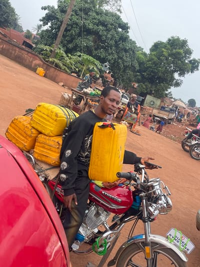 A man in Otukpa, Nigeria, is seated on a red and chrome Kymstone motorcycle. He is holding a smartphone in his left hand. Strapped to the motorcycle are four yellow containers. The scene appears to be a market area. Other motorbikes and vehicles are visible in the background. Trees and buildings are also present in the background. A red vehicle is in the foreground partially obscuring the motorcycle.