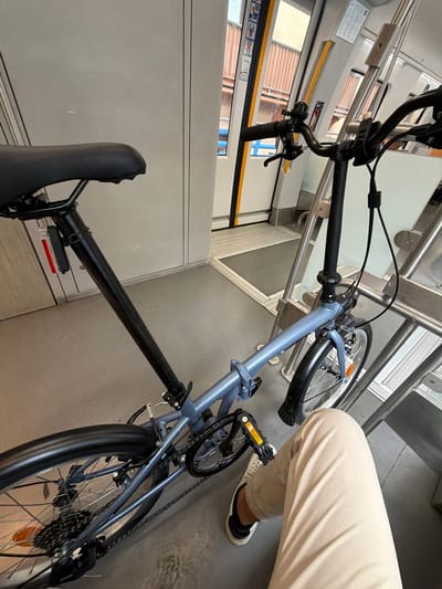 The image shows a light blue/grey folding bicycle stowed inside a public transport vehicle, likely a train or tram, in Pasaia, Spain. The bicycle, which appears compact due to its folding design, rests on a grey floor against light-colored interior walls and metallic railings.

A person's lower body, specifically their legs and one foot, is visible in the foreground, dressed in light-colored trousers and a black and white shoe. They appear to be seated or standing close to the bicycle.

Notable details of the bicycle include black handlebars with grips and brake levers, black foldable pedals with orange reflectors, a black seat, and a black seat post with a small rectangular red reflector. The wheels have black tires and silver spokes, and the rear wheel clearly displays the gear cassette and chain mechanism. The surrounding environment features windows through which blurred outdoor scenery can be observed, characteristic of urban or suburban travel. No legible text or specific branding is visible on the bicycle or within the transport vehicle's interior. The scene suggests a passenger traveling with their bicycle.