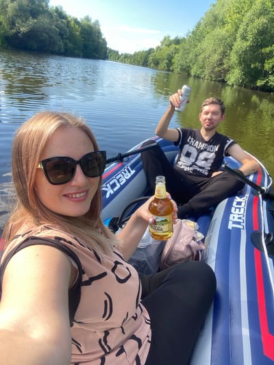 Two people, a man and a woman, are in an inflatable boat on a body of water. The man is wearing a black t-shirt with "CHAMPION 88" printed on it in white. He is holding a silver can in his left hand and smiling towards the camera. The woman is wearing a pink patterned top and sunglasses. She is holding a bottle of beer with a yellow label in her right hand and smiling.

The boat is blue and white with red stripes. The word "TRECK" is visible on the side of the boat. The water is calm and reflects the sunlight, and lush green trees line the shore. The sky is clear and blue, suggesting a sunny day.

The activity appears to be a leisurely outing on the water, possibly a picnic or a relaxing trip in the boat. The overall mood is cheerful and relaxed, with both individuals smiling and enjoying their time. The visible text on the man's shirt and the boat, as well as the beer bottle label, are notable details. The location context is Chulki-Sokolovo, Russia.