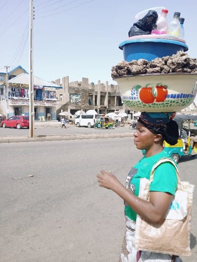 The scene depicts a sunny, urban street in Mumbwa, Zambia. A woman, seen in profile, walks from right to left across the foreground on an asphalt road. She has a dark complexion, wears a black headscarf, a green t-shirt with a white graphic, and a white skirt with brown patterns. A tan shoulder bag hangs from her right shoulder. She is balancing a stack of containers on her head: a large cream-colored bowl adorned with red apples, filled with dark, granular material, topped by a blue plastic basin, which in turn holds several plastic bottles and a black plastic bag. Her right hand is slightly raised, open-palmed.
In the mid-ground, a light-colored utility pole with power lines stands vertically on the left. Behind it is a two-story building with a light blue and white facade, displaying clothing on mannequins on both levels; a sign on the ground floor is partially visible, reading "JANDESO BUILDING". A red sedan is parked in front of this building. To the right, a large, multi-story concrete structure, appearing either unfinished or dilapidated, features exposed framework and multiple open windows, with a large poster on its facade. On the street between these buildings, a white van is parked, alongside several auto-rickshaws (one prominent green and yellow) and other indistinct vehicles. Multiple human figures are sparsely distributed in the background, walking or standing near the vehicles and buildings. The sky is clear and light blue, indicating bright daylight.