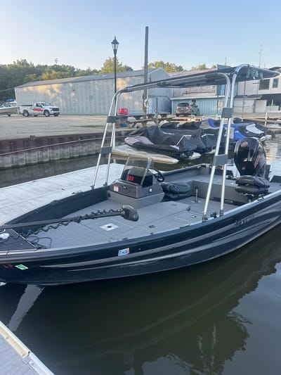 An exterior, eye-level, medium shot shows a dark blue fishing boat docked at a marina on a bright, sunny day. The boat is positioned facing to the right, with its bow to the left of the frame. The boat has a dark gray interior with two bucket seats, a steering console, and a trolling motor mounted on the bow. A Bimini top with a dark gray fabric is extended over the cockpit area, supported by a silver metal frame. Several jet skis covered with dark gray tarps are docked to the right of the fishing boat. In the background, a U-Haul pickup truck is parked near a large, light-colored industrial building with a gray metal exterior. A street lamp with a black lantern is visible behind the truck. Trees line the horizon behind the building. The water in the marina is calm with slight reflections. The scene is located in the city of Algonquin, United States of America.