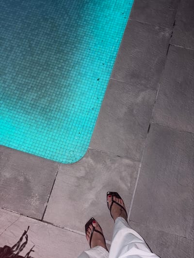 The image shows a close-up, overhead view of a person's feet wearing black thong sandals with red nail polish, standing on a stone tiled patio next to a turquoise tiled swimming pool. The pool is illuminated from within, casting a bright blue-green glow. The patio tiles are large, rectangular, and dark gray with visible grout lines. The person is wearing white pants that extend down to their ankles. A shadow of what appears to be fringed material is cast on the tiles to the left of the person's feet. The scene is likely outdoors, in a domestic or commercial setting, given the presence of a swimming pool and patio. The overall lighting suggests it may be evening or nighttime. The location is identified as Urbanización Encinares De Alcolea, Spain.