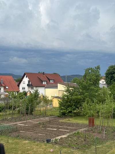 This is a vertical, eye-level outdoor shot taken on a cloudy day in Schömberg, Germany. In the foreground, a neatly maintained vegetable garden is visible, with rows of young plants and wooden stakes. A rusty barrel stands amongst the plants, and a black bucket is on the grass. A chain-link fence borders the garden. Behind the garden, a residential area is depicted with houses featuring red-tiled roofs. Some of the houses have visible windows and balconies. Lush green trees and bushes are scattered throughout the scene. In the background, rolling hills and distant power lines are visible under a dramatic, overcast sky. The overall impression is that of a peaceful, rural suburban environment.
