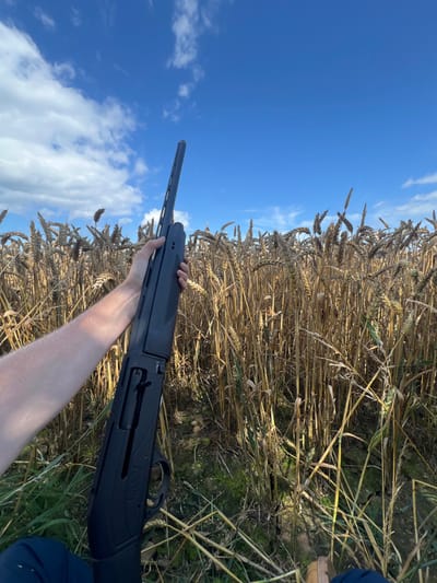 The image captures a field of mature, dry, golden-brown wheat stalks under a clear blue sky with scattered white clouds, in Skipsea, United Kingdom. A person's fair-skinned right forearm and hand are visible in the lower left, holding a dark-colored semi-automatic shotgun, identified by the "ESCORT" brand name visible on its receiver. The shotgun's barrel is angled upwards and to the right, pointing towards the sky. The action of the shotgun is open, exposing internal mechanical components. Portions of dark blue clothing are visible near the bottom left, and a light-colored boot or shoe is partially visible at the bottom right. The ground beneath the wheat stalks contains patches of green vegetation and several light-colored, spherical objects.