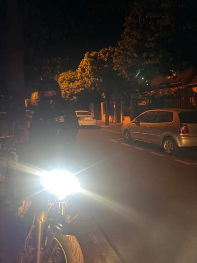 At night in Joinville-le-Pont, France, a motorcycle with its headlight on is parked on the side of the road next to a parked silver car. A person is on the motorcycle. Trees are visible in the background.