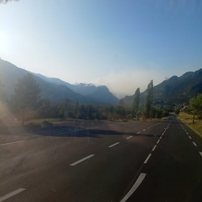 The image depicts a scenic view of a road in the vicinity of Saint-Crépin, France, likely taken during daytime. The foreground features an asphalt road with clear white lane markings. To the left of the road, there is a grassy area and a few trees, with a prominent pine tree casting a shadow. In the middle ground, a dense forest stretches across the landscape, with a visible layer of haze or smoke rising from behind the trees and obscuring the lower portions of the mountains. The background is dominated by a range of imposing, hazy mountains under a clear blue sky. A few scattered structures, possibly houses or buildings, are visible on the right side of the image, nestled amongst the trees and fields at the foot of the mountains. The overall impression is one of a rural, mountainous area.