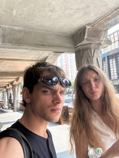 The image captures two individuals in a selfie, likely taken in the Hongkou District of Shanghai, China, based on the presence of signage in Chinese characters. The subjects appear to be a young man and woman. The man is in the foreground, wearing sunglasses perched on his head, a dark-colored shirt, and carrying a backpack strap. The woman, with long blonde hair, is behind him, holding a bottle of water. The background features architectural details, including concrete structures, possibly part of a building or historical site, with signs indicating the urban environment. The overall scene suggests a casual outing or tourist activity.