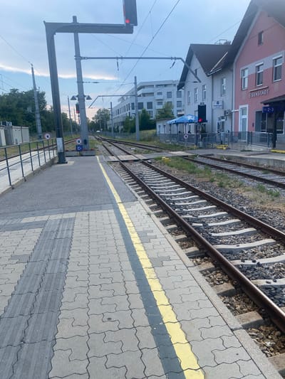The image captures a view of the train station in Eisenstadt, Austria. The foreground features a train platform with patterned paving stones and a yellow safety line. Adjacent to the platform are railway tracks. In the background, a pink building with "EISENSTADT" signage is visible, along with other buildings and trees. Overhead electrical lines for the trains are prominent. A red train signal is illuminated. The sky appears overcast with some clouds. There are no visible people actively engaged in the scene. The overall environment depicts a functional train station setting.