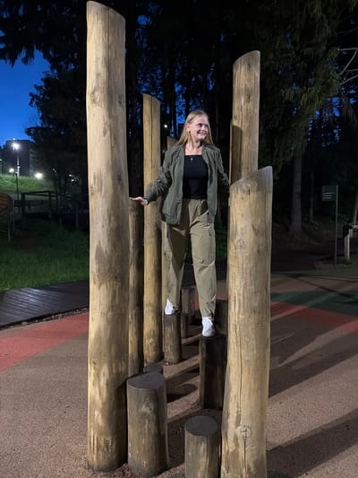 A young woman with blonde hair is standing on a wooden obstacle course in a park at dusk. She is wearing a dark t-shirt, a green jacket, and olive-colored cargo pants. She is positioned between four large wooden posts, with her left hand resting on one of the posts. In the background, there are trees, lights, and a sign with Cyrillic text, suggesting the location is in Izhevsk, Russia. The ground is covered with a dark, textured material.