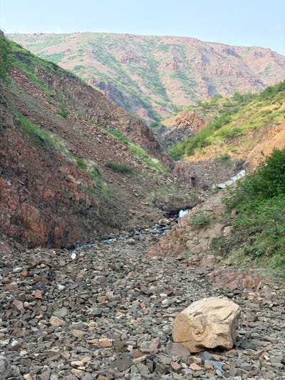 The image shows a rocky, mountainous landscape with a narrow riverbed running through a canyon. The slopes are covered with rocks and sparse vegetation, predominantly green grasses and shrubs. The riverbed itself is also filled with stones of various sizes. In the foreground, a large, light-colored boulder rests on a bed of smaller rocks. The canyon walls are steep and composed of reddish-brown rock formations. In the distance, the mountains recede, their peaks softened by a light haze. The sky above is a pale, clear blue. This scene is located in the vicinity of Talnakh, Russia, an industrial city in Krasnoyarsk Krai, known for its mineral deposits and surrounding natural beauty.