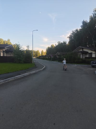 The image shows a street in the city of Krasnoe Pole, Russia, with a boy riding an electric scooter on the asphalt road. The boy is wearing a light-colored t-shirt, shorts, and a cap. Houses with pitched roofs and trees line the street. A street lamp is visible on the left side of the road. The sky is clear with a crescent moon and some wispy clouds. A dark-colored car with a license plate is parked on the right side of the road. The road has a sidewalk on the left, separated by a curb. The area appears to be a residential neighborhood.