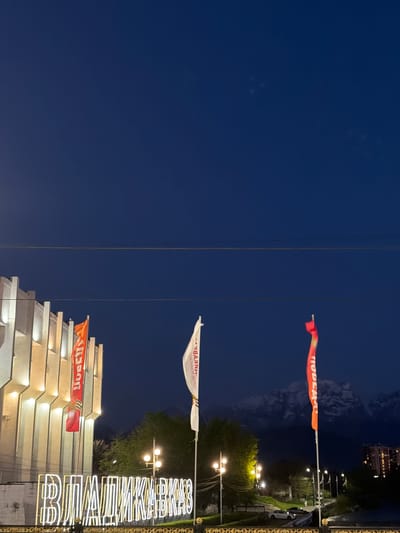 The image shows a nighttime scene with several flags and illuminated signs in the city of Chermen, Russia. In the top-right corner, an orange flag with the word "ПОБЕДА" (Victory) is displayed. Below that, a white flag is seen with the words "СЛАВА ВОИНАМ ДОНСКОЙ ЗЕМЛИ" (Glory to the Warriors of the Don Land). A building with orange flags is located in the lower part of the image, and the sign "ВЕЛИКИМ КИМКОВЦАМ" (To the Great KIM workers) is vertically displayed. Streetlights are visible. The background shows a dark blue sky and outlines of distant mountains. Powerlines run vertically across the image.