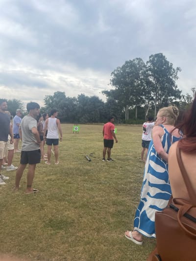 People stand in a grassy field facing a green target. The group consists of men and women wearing casual clothing like shorts, t-shirts, and a blue patterned dress. One person is wearing a red shirt. The background includes trees and a cloudy sky. A small speaker and a quiver are on the grass near the target.