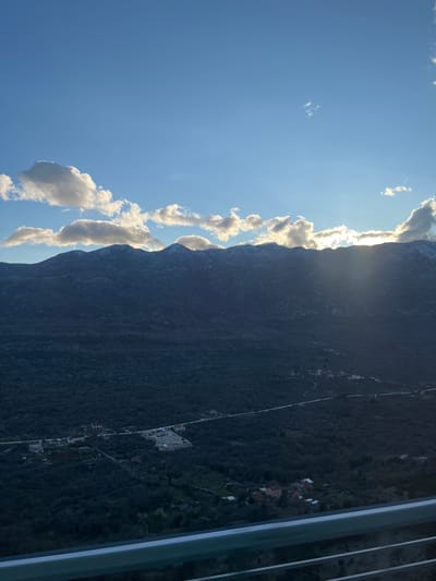 The image shows a mountain landscape with a partially clouded sky. The sun shines through a gap between the clouds and the mountain peak. The mountain appears dark and rugged. A settlement, Rašćane Gornje, Croatia, with houses and a road, is visible on the mountain's right-hand slope. Steel beams are present in the right foreground, suggestive of a bridge or structure.