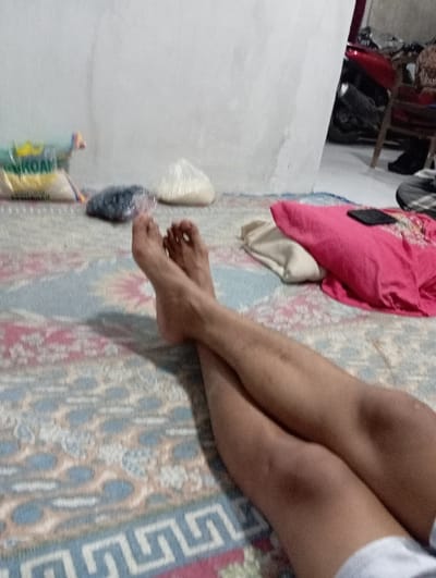 The image depicts an indoor scene in Bulukandang, Indonesia. The foreground is dominated by the lower legs and bare feet of an individual, positioned with ankles crossed, resting on a patterned floor rug. The individual wears white shorts, partially visible at the bottom right. The rug features a repeating geometric design in shades of blue, white, pink, and beige. In the mid-ground on the rug, from left to right, are a large white sack with green and yellow markings (partially labeled "KOAM"), a dark-colored bag wrapped in clear plastic, and a clear plastic bag containing white granular material. To the right, on the rug, a bright pink cushion or folded fabric item is present, with a dark-colored smartphone resting on its surface. Beneath or beside the pink item is a folded light-colored, possibly beige, fabric item. The background consists of a plain, light-colored wall. In the upper right quadrant, a red motorcycle and a portion of a wooden chair are visible on a lighter floor surface beyond the rug.