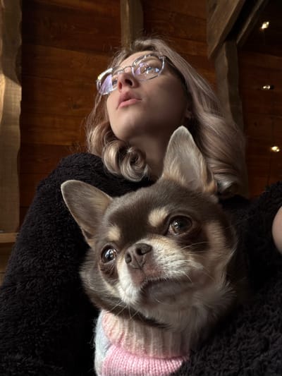 A female individual is captured indoors in Доња Брезна, Montenegro, holding a small brown and tan Chihuahua dog. The female has blonde, wavy hair, wears clear-framed glasses, and is dressed in a dark, textured garment. Her head is tilted upwards and to the left, with her gaze directed towards the upper left portion of the frame. Her lips are slightly parted. The Chihuahua, wearing a pink, white, and blue striped knitted sweater, is positioned directly in front of the female, resting against her chest. The dog has large, erect ears and dark eyes, and its gaze is directed forward and slightly upwards. The background features light brown wooden paneling and a darker vertical wooden beam on the right. Small, illuminated string lights are visible in the upper right background.