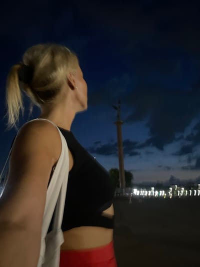 A blonde woman is captured in a close-up shot, from the shoulders up, as she looks to her right. She wears a black top and red bottoms, with a white bag slung over her left shoulder. Her hair is pulled back in a ponytail, and she has a small hoop earring in her visible ear. In the background, a tall monument stands against a dark, cloudy sky. A blurred line of lights stretches across the horizon, suggesting a city or town setting. The overall scene is dimly lit, indicating it is either dusk or nighttime. The location is Verkhnyaya Stavropol'ka, Russia.