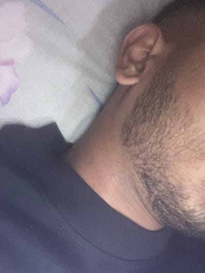 The image captures a close-up, angled view of a person resting indoors, likely in Zetiovo, Bulgaria. The primary subjects are the right side of a person's head, specifically their ear, jawline, and neck, partially covered with stubble or a short beard. The person is lying on a pillow, which has a light-colored cover featuring faint purple and blue abstract patterns, possibly floral or leafy. A dark blue garment, likely a shirt or jacket, is visible around their neck and shoulder area.
The scene depicts a moment of repose. No distinct activity is occurring beyond the person appearing to be asleep or at rest.
Notable details include the texture of the stubble/beard and the skin on the neck. The image has a grainy quality, suggesting dim lighting conditions. The garment is a solid, dark blue color. No specific time of day or weather conditions are evident from the indoor setting. There are no visible facial expressions to convey emotion, and no text is visible in the frame. The overall impression is one of a quiet, personal, and potentially domestic setting.