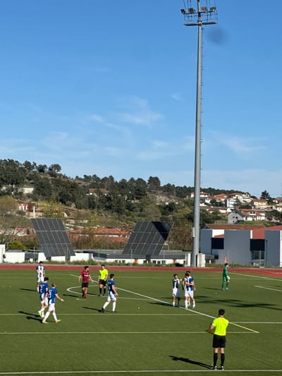 The image depicts a soccer field adjacent to a solar panel installation, located near a hillside residential area possibly near or in Valpaços, Portugal. A light tower extends into a clear blue sky. Several people are on the field, appearing to be players and a referee. The players are wearing uniforms in blue and white, and red and black. The referee is wearing a bright yellow shirt. The scene suggests a soccer game or practice is in progress. The solar panels are adjacent to the field, near a building. A neighborhood of houses and buildings is seen on a hill in the background.