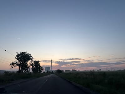 The image presents a rural landscape scene at either sunrise or sunset, looking down a two-lane asphalt road that recedes into the distance. The sky dominates the upper portion, displaying a serene gradient from a pale blue at the top to soft hues of orange, pink, and yellow along the horizon, indicating the low position of the sun. A single, thin white contrail from an aircraft is visible in the upper-left part of the sky.
On the left side of the road, a cluster of dark, silhouetted trees and bushes rises against the luminous horizon. Overhead, multiple thin power lines extend across the frame. Further down the road on the left, a prominent industrial complex, likely a thermal power plant, stands out with its very tall, slender chimney and blocky buildings.
To the right of the road and extending into the background are expansive fields covered in grass and scattered with smaller trees. A faint mist or fog lies low over these distant fields, particularly on the right, suggesting the coolness of early morning or late evening. Several electricity pylons are discernible in the hazy distance on the right, connecting to the power lines seen overhead. The road itself appears dark and somewhat reflective.
No people or explicit text are visible in the image. The setting suggests a landscape characteristic of the area around Obruchishte, Bulgaria, with a blend of natural scenery and industrial infrastructure.