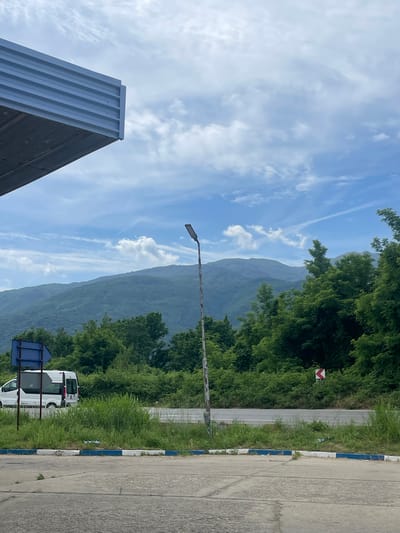 This image, captured in Parvomai, Bulgaria, presents a daytime view of a mountainous landscape alongside a road. The scene is dominated by a clear blue sky interspersed with white, wispy clouds and a visible contrail from an aircraft.
In the foreground, on the left side, is the corner of a modern building featuring a corrugated, light-colored facade. A tall, dark lamppost stands vertically in the mid-ground, positioned centrally, with its light fixture pointing towards the left.
Behind the lamppost and extending across the background are large, lush green mountains, their slopes covered in dense vegetation, appearing somewhat hazy due to distance. On the right side of the image, thick green trees and bushes line a paved road or parking area. This road features a distinctive blue and white painted curb along its edge. Further down the road, a red and white chevron sign, indicative of a sharp turn or hazard, is visible among the greenery. A white van is partially visible, parked near a blue information sign post on the right side of the road. The weather appears fair and bright. No discernible text is visible on any signs or objects.