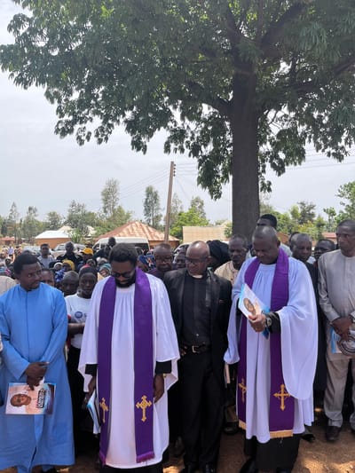 The image shows a procession of people, primarily clergy, in an outdoor setting that is possibly the city of Dogon Dinia, Nigeria. The group of clergymen are wearing traditional white robes with purple stoles adorned with gold crosses. Some individuals are holding what appear to be funeral programs featuring a portrait of a man named "Dowson Baba Akut". In the background, there is a residential area with trees, power lines and a corrugated iron roof visible. A large tree dominates the left side of the image. The ground appears to be dirt.