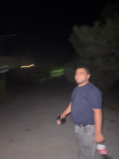 In the city of Egorlykskaya, Russia, at night, a man is captured in a motion-blurred image. He is walking on a dirt path, possibly in a rural area or park, with trees visible in the background. The man is wearing a dark blue t-shirt and gray pants, with a black belt. He is smiling and looking to his right. He is holding a dark object in his left hand, and a red and white bottle in his right hand. The scene is dimly lit, suggesting it is nighttime. The motion blur indicates movement, possibly the photographer or the subject walking.