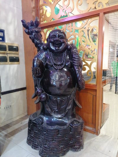 The image shows a statue of a laughing Buddha, positioned indoors. The statue is made of a dark, polished material. It is wearing a beaded necklace. The Buddha holds a gourd in one hand and a staff with decorative elements in the other. The statue stands on a tiered base. Behind the statue is a wooden partition with decorative cutouts. A person is standing behind a beaded curtain in the background. The location is Chu Văn An Ward, Vietnam.
