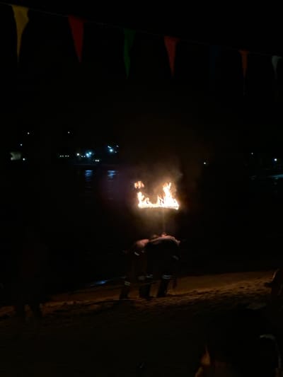 The image captures a nighttime scene along the waterfront of Hat Khai Muk, Thailand. In the foreground, a person is seen blowing fire toward a burning structure or art piece, possibly for entertainment or a ritualistic performance. The scene is lit primarily by the fire and reflected by the water. A few people stand nearby and watch. Along the far side of the water, there are strings of small lights, likely marking the edge of a structure, building, or path. Banners are seen on the left side of the image.