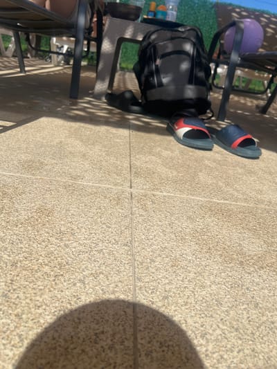 A pair of blue and red slides rests on a textured beige tiled patio floor. Behind the slides, a gray and black backpack is positioned next to a chair. In the background, another chair with a purple ball on it is visible. The scene is lit by sunlight casting shadows on the patio. The location is identified as Delchevo, North Macedonia.