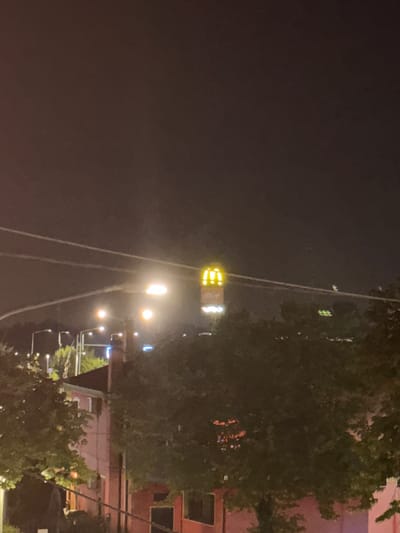 The image depicts a nighttime scene in the city of Spinea, Italy, featuring a lit McDonald's sign as the central focal point. The sign, illuminated in yellow, displays the iconic golden arches and the text "McCafé" below. It is mounted on a tall pole, suggesting a roadside location. In the foreground, obscured by trees, is a building with a pinkish hue and visible windows, possibly residential or commercial. The environment is characterized by streetlights casting a glow on the surroundings, and power lines crisscrossing the frame. The overall atmosphere is dimly lit, typical of an urban night.