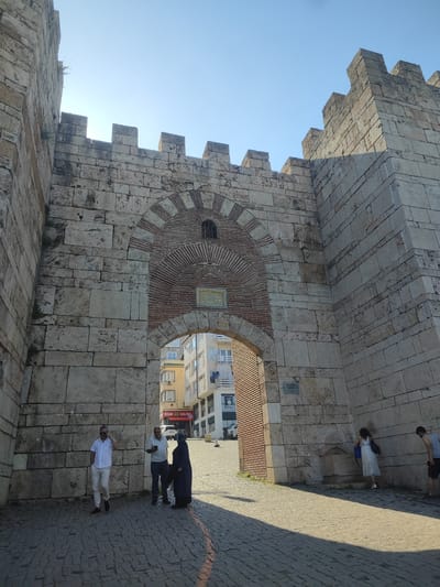 The image depicts a stone gateway in Osmangazi, Turkey, under a clear blue sky, suggesting a sunny day. The gateway, constructed from large, weathered stone blocks and accented with brickwork in its arched opening, appears ancient and imposing. The tops of the walls are crenellated, indicative of a defensive structure.

Several people are visible near the gateway. On the left, a man in a white shirt and light-colored pants, wearing sunglasses, is talking on his phone while walking. To his right, two individuals stand together, one in a light-colored shirt and the other in dark clothing, possibly a hijab. Further to the right, a woman in a white dress is drinking from a stone fountain built into the wall, while a man in a grey tank top and shorts stands nearby.

The scene suggests a casual, daytime activity, with people passing through or resting near the historical structure. In the background, modern buildings and a street are visible, indicating that this ancient gateway is integrated into a contemporary urban environment.

A small plaque is mounted on the stone wall to the right of the fountain, bearing the text "SULTANAH NAPIŞI" and smaller script below it. Another sign is visible on the building in the background, with the word "SALON" prominently displayed. The overall impression is one of historical significance meeting everyday life.