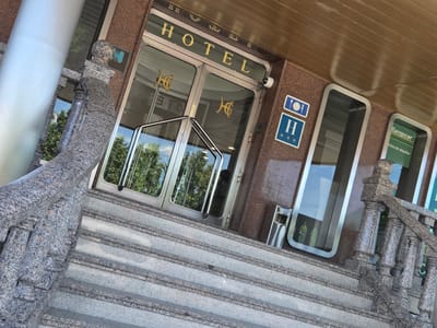 The image shows the entrance to a hotel and a Europcar rental agency. The hotel entrance features double glass doors with the word "HOTEL" in gold lettering above them. Two hotel logos, appearing to be stylized "HG" or "GH", are visible on the glass doors. Below the "HOTEL" sign, there are two blue and white signs: one with a fork and knife icon and a "H" with three stars, indicating a hotel. Adjacent to the hotel entrance, to the right, is a Europcar rental agency with a green sign featuring the Europcar logo and text in Spanish that translates to "rental office". The entrance is framed by granite-clad walls and a polished granite balustrade and staircase. The scene is illuminated by natural daylight. A silver trash receptacle is located to the right of the hotel entrance. The location is identified as La Estación, Spain.