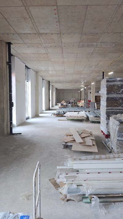 This is a full-frame, eye-level, vertical shot of the interior of a building under construction. The scene is located within a large, open space with a concrete floor and ceiling. The ceiling is composed of square concrete panels with visible seams and red and blue markings, presumably for construction purposes. Along the left wall, a series of large windows are present, separated by concrete columns. Black pipes are also visible along this wall.
The space is filled with various construction materials and equipment. On the right side of the image, large stacks of materials wrapped in plastic are piled high. In the foreground and middle ground, there are piles of wooden planks, metal scaffolding components, and other building supplies. A metal hand truck with white handles is visible in the bottom left of the frame. The overall impression is one of an unfinished interior space with materials organized for future work. No people are visible in the image. The lighting appears to be a mix of natural light from the windows and artificial overhead lighting. The location is a construction site in Oxford, United Kingdom.